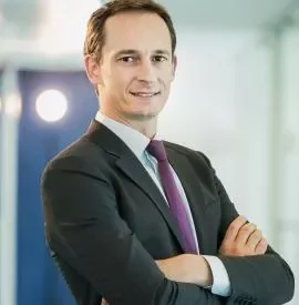 Man in a suit smiling with arms crossed in an office setting.