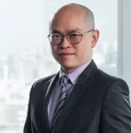 Man in a suit with glasses standing in front of a window with a city view.