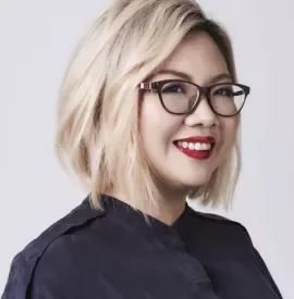 Smiling person with blonde hair and glasses wearing a dark blouse.
