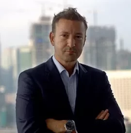 Man in a suit standing with arms crossed in an office with a cityscape background.