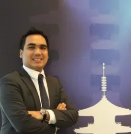 Smiling person in a suit standing with arms crossed, blue background with white pagoda design.