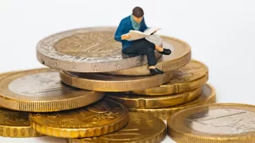 Miniature figure reading a book while sitting on a stack of coins.