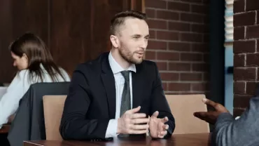 Two businessmen in conversation at a cafe, with two women using laptops in the background.