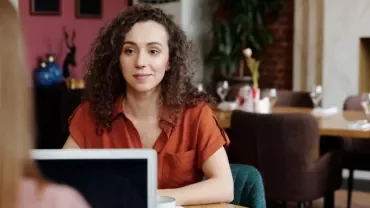 Woman with curly hair in a cafe, facing another person with a laptop.