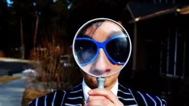 Person in striped suit holding magnifying glass over blue sunglasses.