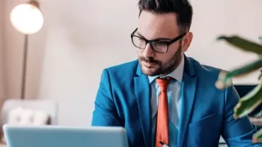 Man in a blue suit and red tie working on a laptop at a desk.