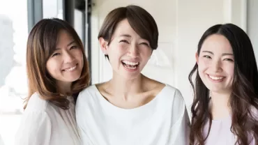 Three women smiling indoors near a window with city view.