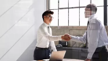 Two men in a bright office shaking hands near a laptop.