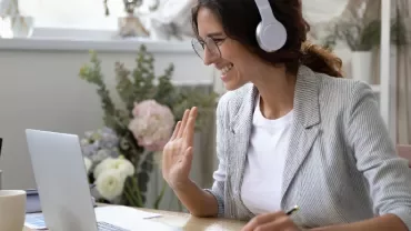 Woman in headphones waving during a video call on a laptop.