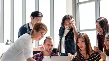 A diverse group of people excitedly looking at a laptop screen in a bright office.