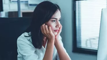 Woman in a white shirt looking thoughtfully at a computer screen in an office.