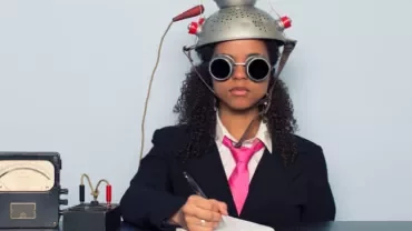 Person in a suit with goggles and a colander on head writing at a desk with vintage equipment.