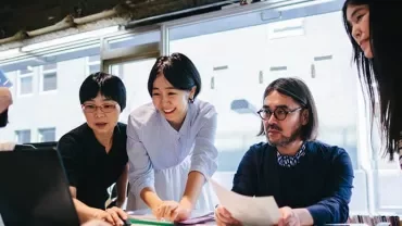 Group of people gathered around a laptop in an office, discussing ideas.