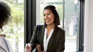 Two women shaking hands by a window, smiling at each other.