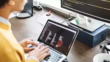 Person coding on a laptop with monitor showing code.