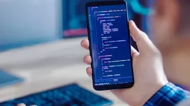Person holding a smartphone displaying code, sitting at a desk with a computer monitor in the background.