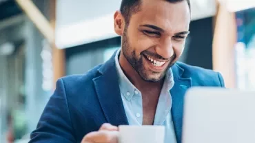 Man smiling while using a laptop in a cafe, holding a cup of coffee.