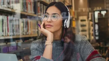Woman in a library wearing headphones, gazing thoughtfully.