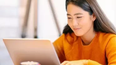 Woman in orange sweater using a laptop at a desk.