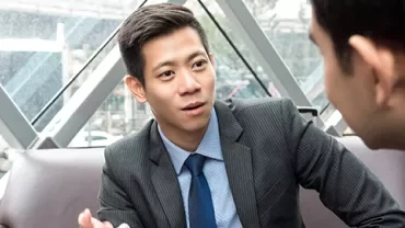 Two people in a business meeting, one speaking, seated near large windows.