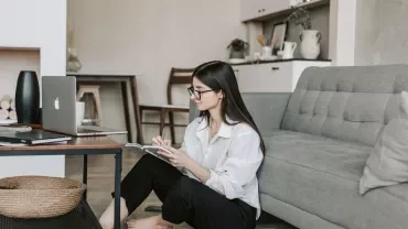 Woman reading a book on the floor near a laptop in a cozy living room.
