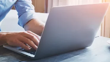 Person typing on a laptop next to a cup on a wooden table in sunlight.