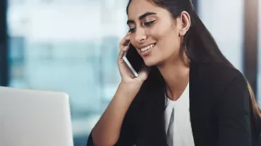 Woman smiling while talking on the phone and using a laptop in an office setting.