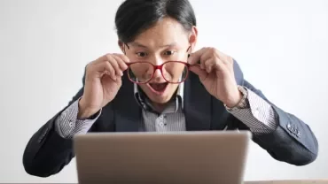 Man looking surprised while holding glasses and staring at a laptop.