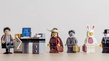 LEGO figures dressed in various costumes stand in line beside an office desk with a computer.