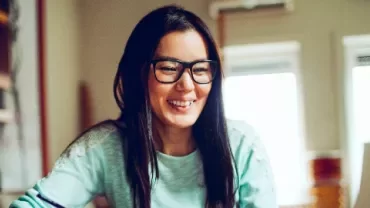 A woman with glasses smiling while using a laptop at home.