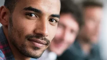 Man in a checkered shirt smiles, with blurred people in the background.
