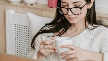 Person wearing glasses, sitting on a bed with a cup, and using a laptop.