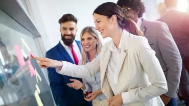 Smiling business professionals with good communication skills at a modern office, with a woman in a white suit pointing at a digital screen while colleagues look on.