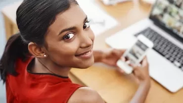 A female HR assistant sitting in front of an open laptop smiling and holding a mobile phone.
