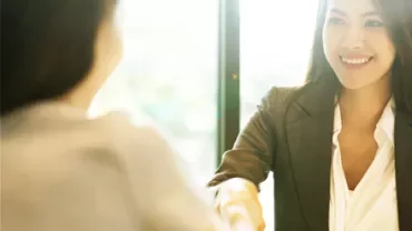 Two women shaking hands in a brightly lit office, smiling at each other.