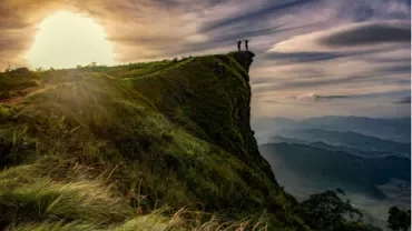 Two people stand on a cliff edge at sunrise, overlooking a vast mountainous landscape.