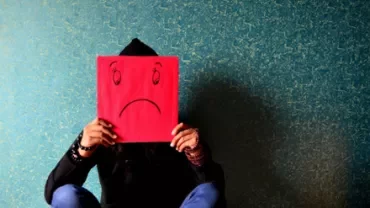 Person sitting against a wall holding a red sad face drawing over their head.