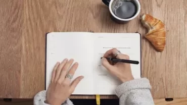 Person writing in a notebook with a coffee and croissant on a wooden table.