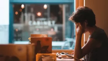 Person sitting in a cafe by a window, wearing earphones and holding their head in one hand.