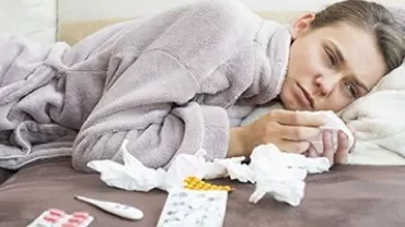 Woman lying on couch with tissues, thermometer, and medicine, looking unwell in a bathrobe.