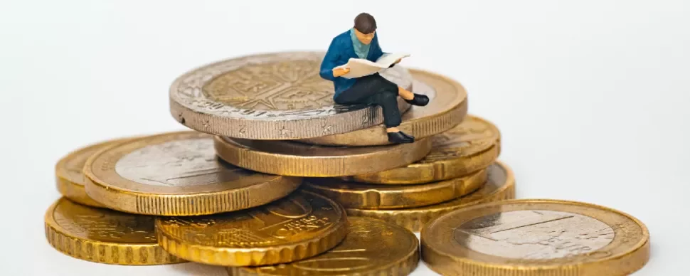 Miniature figure reading a book while sitting on a stack of coins.