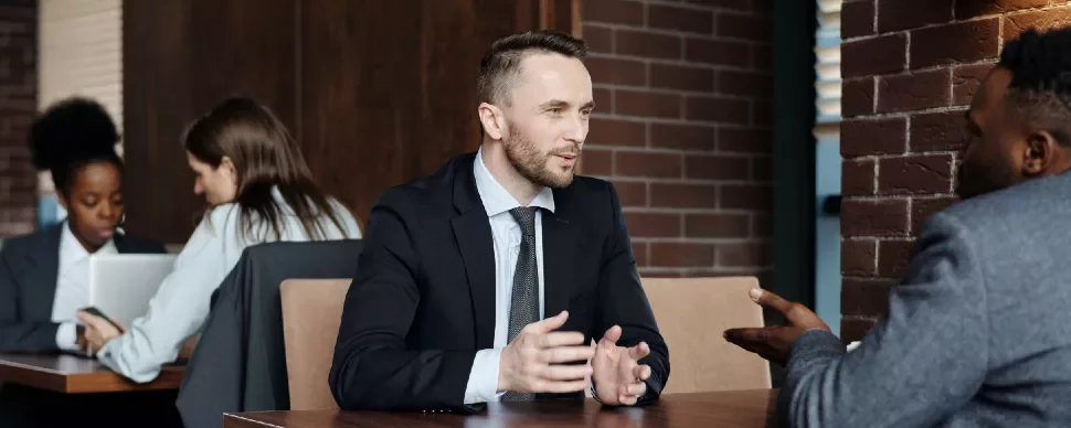 Two businessmen in conversation at a cafe, with two women using laptops in the background.