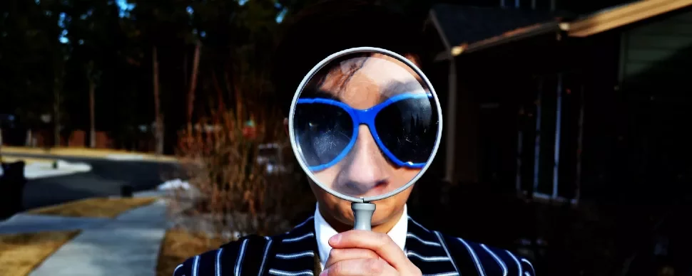 Person in striped suit holding magnifying glass over blue sunglasses.