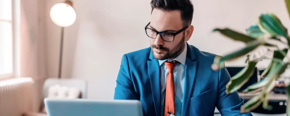 Man in a blue suit and red tie working on a laptop at a desk.