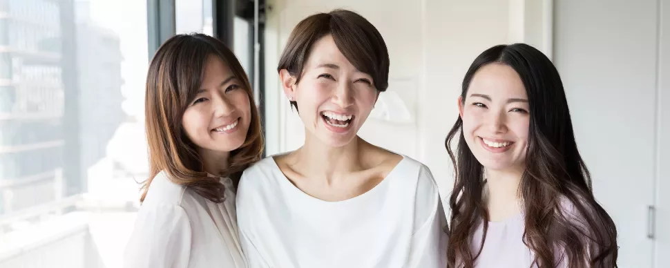 Three women smiling indoors near a window with city view.