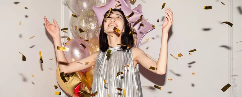 A woman in a shiny dress joyfully throws gold confetti in the air by balloons.