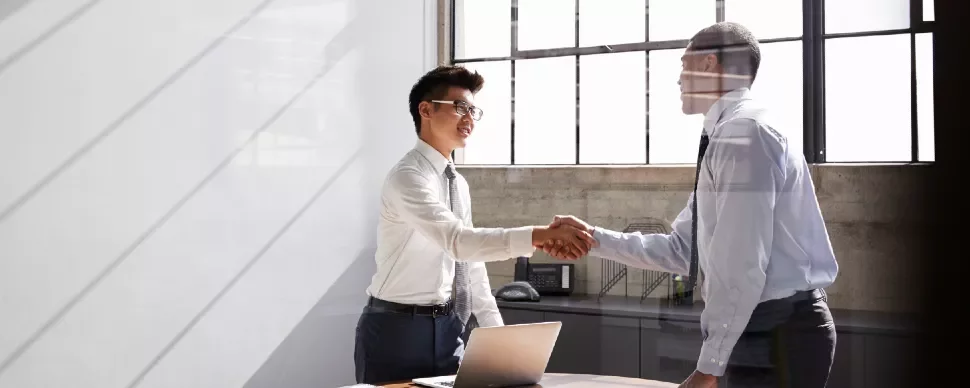 Two men in a bright office shaking hands near a laptop.