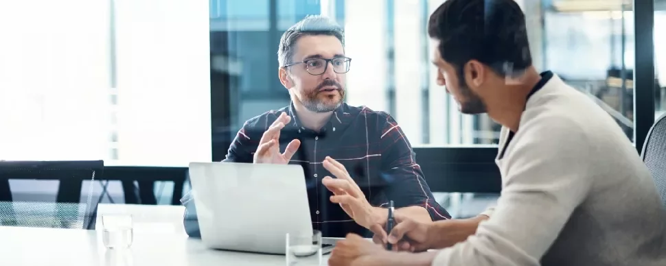 Two men having a discussion in an office with a laptop open on the table.