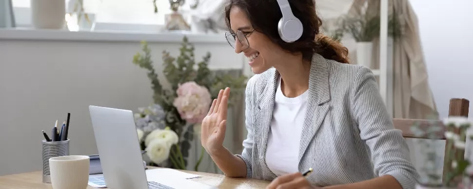 Woman in headphones waving during a video call on a laptop.