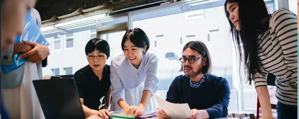 Group of people gathered around a laptop in an office, discussing ideas.